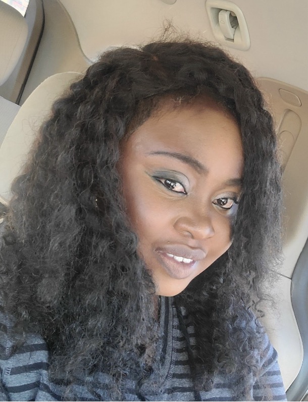 Close-up selfie of a smiling person with long curly hair and subtle eye makeup, sitting in a car.